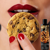A close-up of a person with red lipstick and matching red nail polish holding a chocolate chip cookie near their lips. In the foreground, a bottle labeled "Wet Desserts 'Chocolate Chip Cookie' Warming Lubricant 🍪 | 3oz" is positioned beside the cookie, highlighting its water-soluble warming formula.